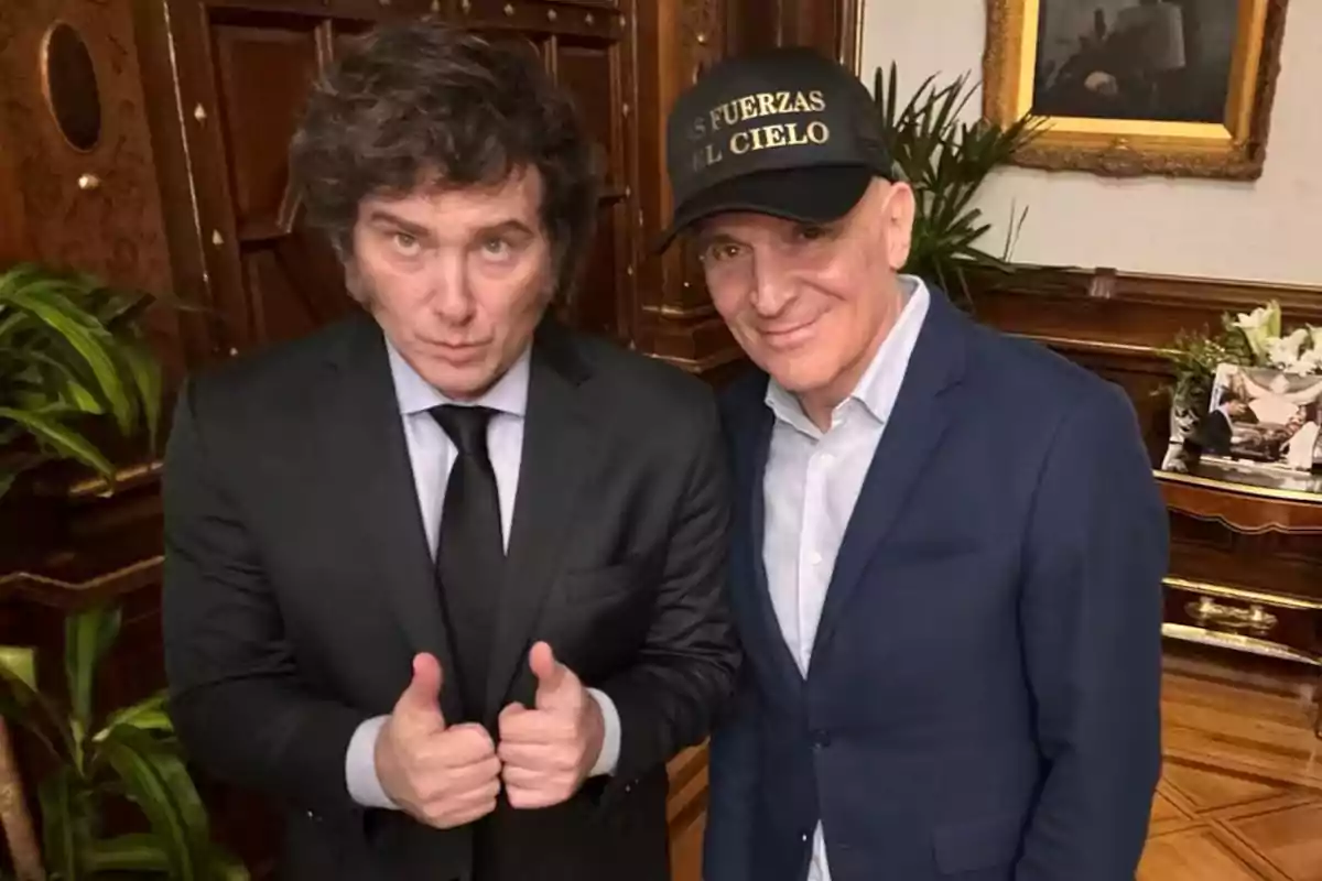 Dos hombres posan juntos en una habitación elegante, uno de ellos lleva traje y corbata y muestra ambos pulgares hacia arriba, el otro viste saco azul y gorra negra con letras doradas