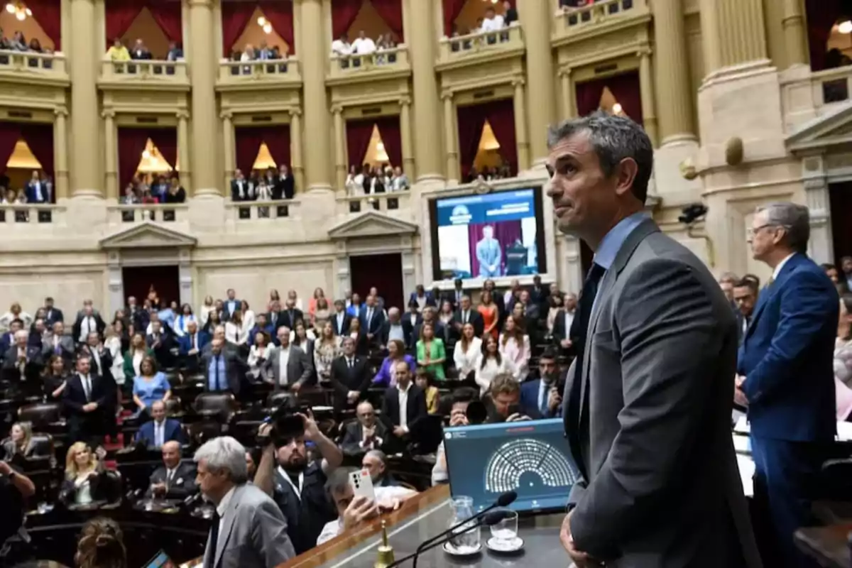 A man in a suit standing in a parliamentary chamber filled with people, with balconies and red curtains in the background. A man in a suit standing in a parliamentary chamber filled with people, with balconies and red curtains in the background.