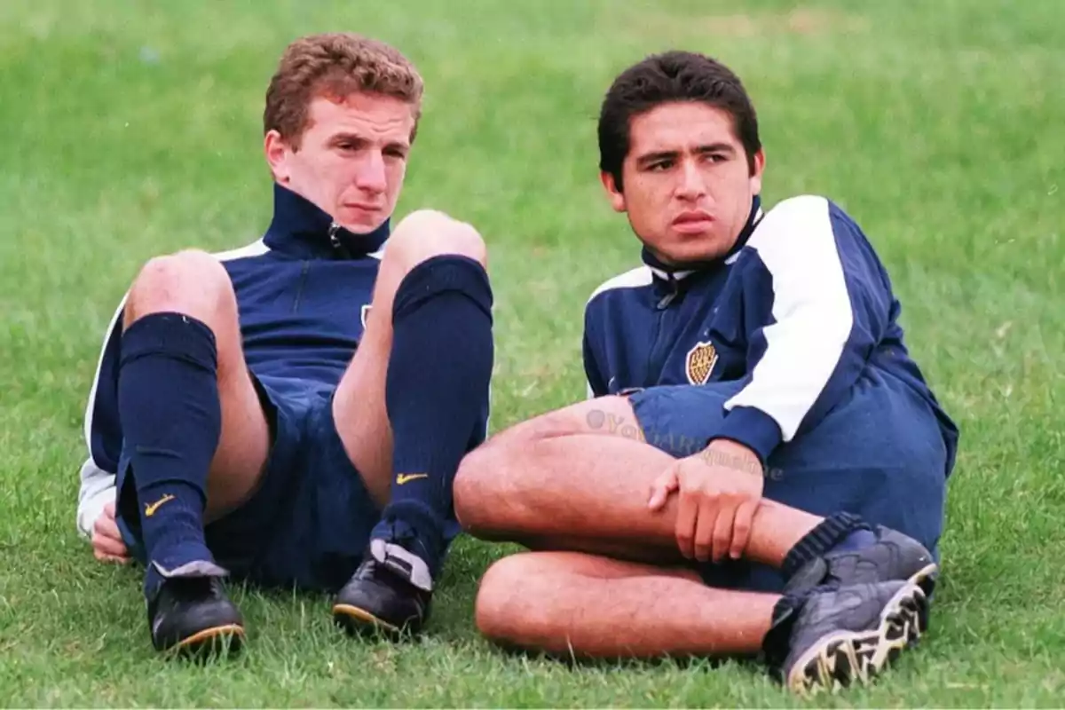 Two players wearing Boca Juniors training gear are sitting on the grass looking to the side. Two players wearing Boca Juniors training gear are sitting on the grass looking to the side.