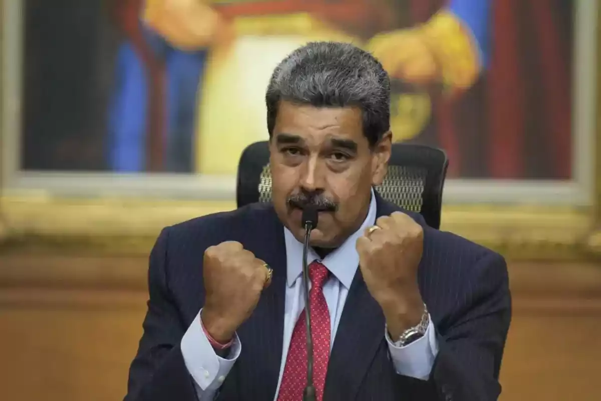 Gray-haired man with a mustache wearing a dark suit and red tie raising both fists in front of a microphone in a room with a painting in the background