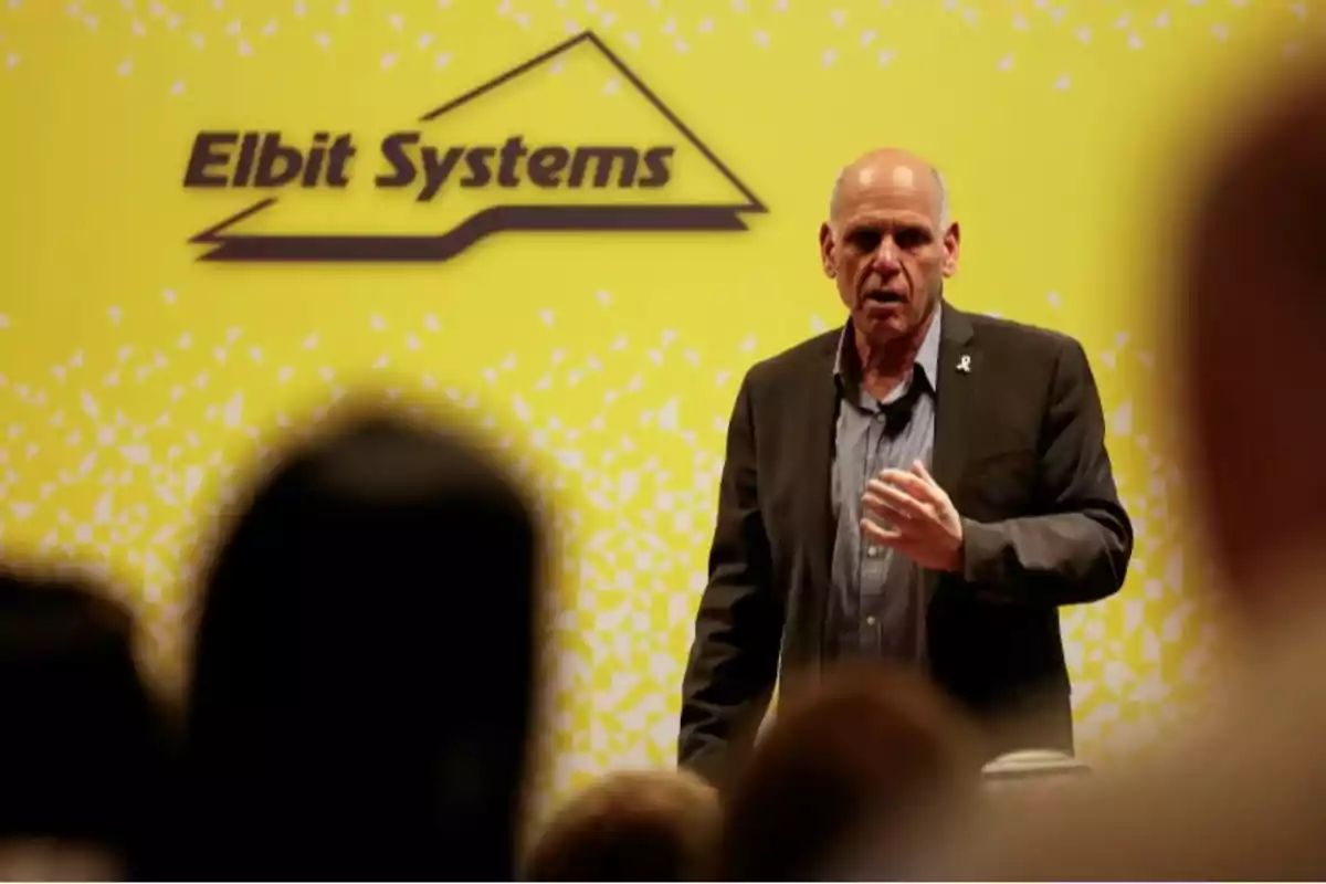 Man standing and speaking in front of an audience with a yellow background featuring the Elbit Systems logo Man standing and speaking in front of an audience with a yellow background featuring the Elbit Systems logo