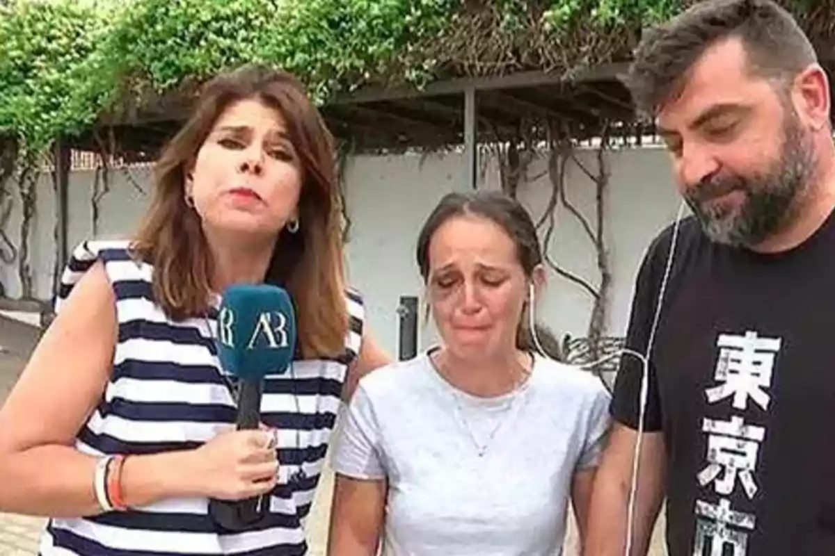 Una reportera sostiene un micrófono mientras entrevista a una mujer visiblemente afectada y a un hombre que la acompaña, todos de pie al aire libre frente a una pared cubierta de plantas. Una reportera sostiene un micrófono mientras entrevista a una mujer visiblemente afectada y a un hombre que la acompaña, todos de pie al aire libre frente a una pared cubierta de plantas.