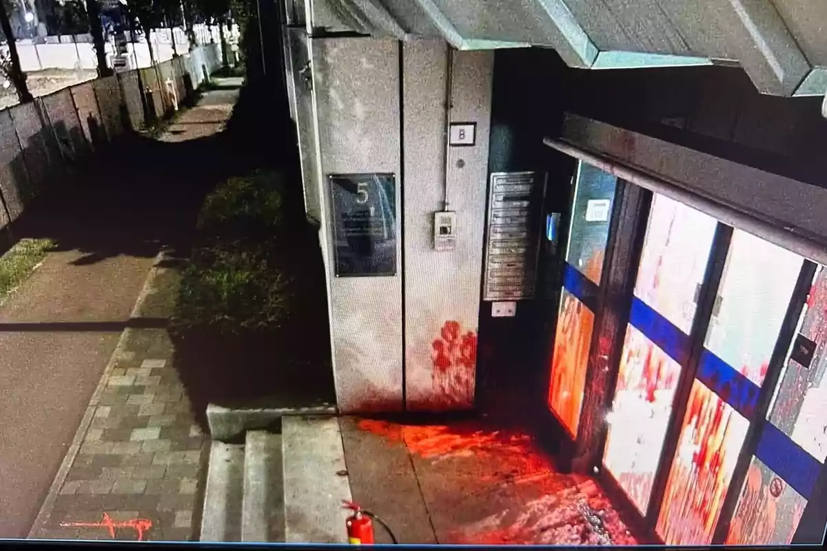 Entrada de un edificio con manchas rojas en la pared y el suelo, junto a una puerta de vidrio y un extintor en la acera