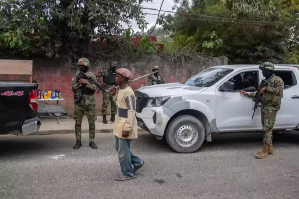 Un hombre camina por la calle mientras varios soldados armados vigilan cerca de una camioneta blanca y una camioneta negra en un entorno urbano Un hombre camina por la calle mientras varios soldados armados vigilan cerca de una camioneta blanca y una camioneta negra en un entorno urbano