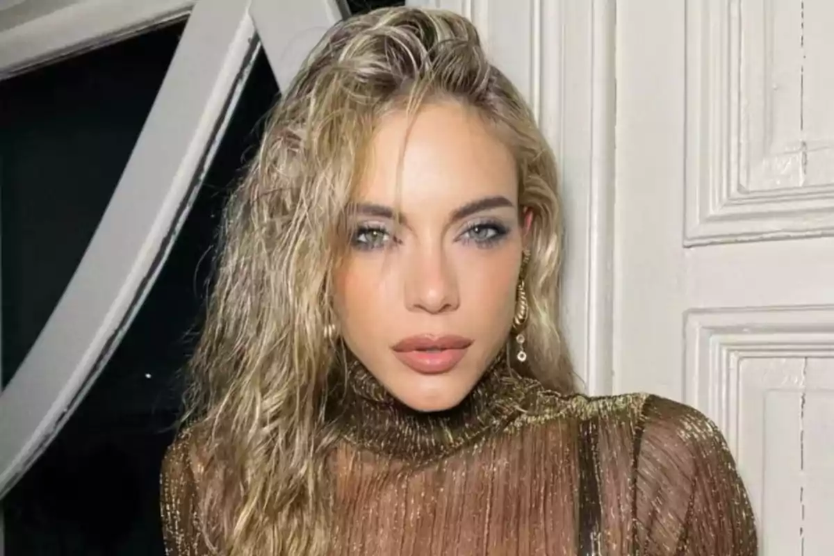 Blonde woman with loose, wavy hair, wearing a shiny gold dress and long earrings, posing in front of a white door