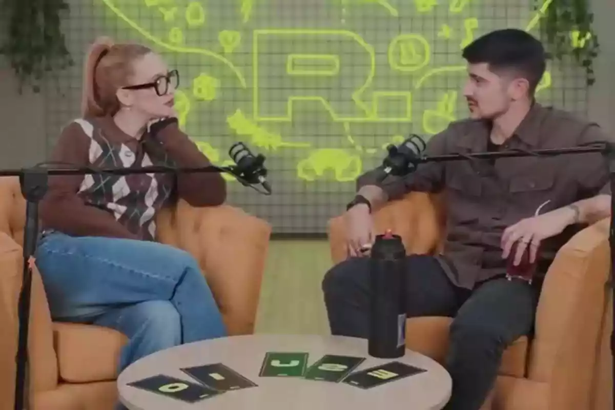 Dos personas conversan sentadas en sillones naranjas frente a micrófonos con una mesa pequeña entre ellas y un fondo con luces y dibujos amarillos Dos personas conversan sentadas en sillones naranjas frente a micrófonos con una mesa pequeña entre ellas y un fondo con luces y dibujos amarillos