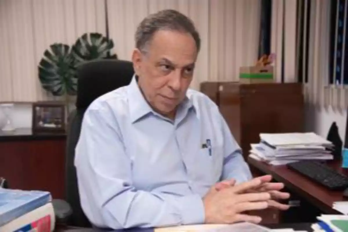 Hombre de cabello canoso y camisa azul claro sentado en una oficina con papeles y una computadora sobre el escritorio