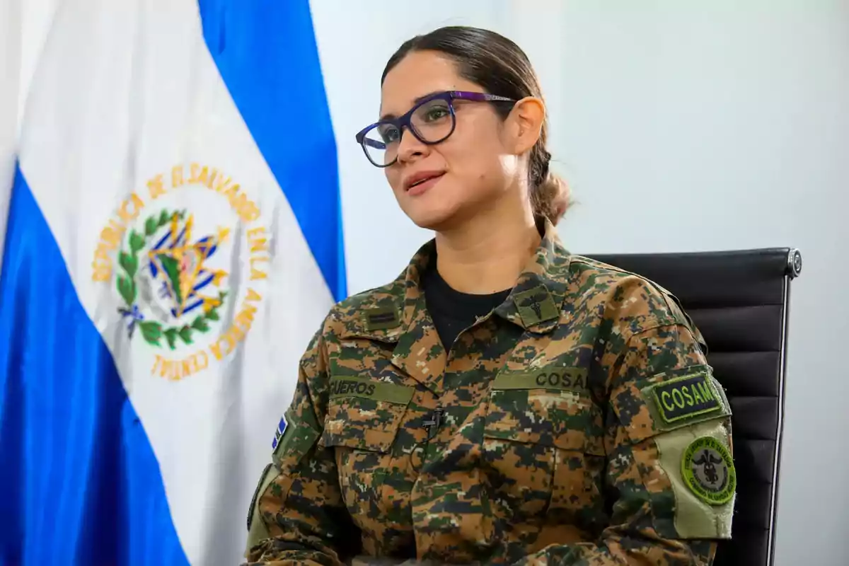Mujer militar con gafas y uniforme de camuflaje sentada frente a una bandera de El Salvador Mujer militar con gafas y uniforme de camuflaje sentada frente a una bandera de El Salvador