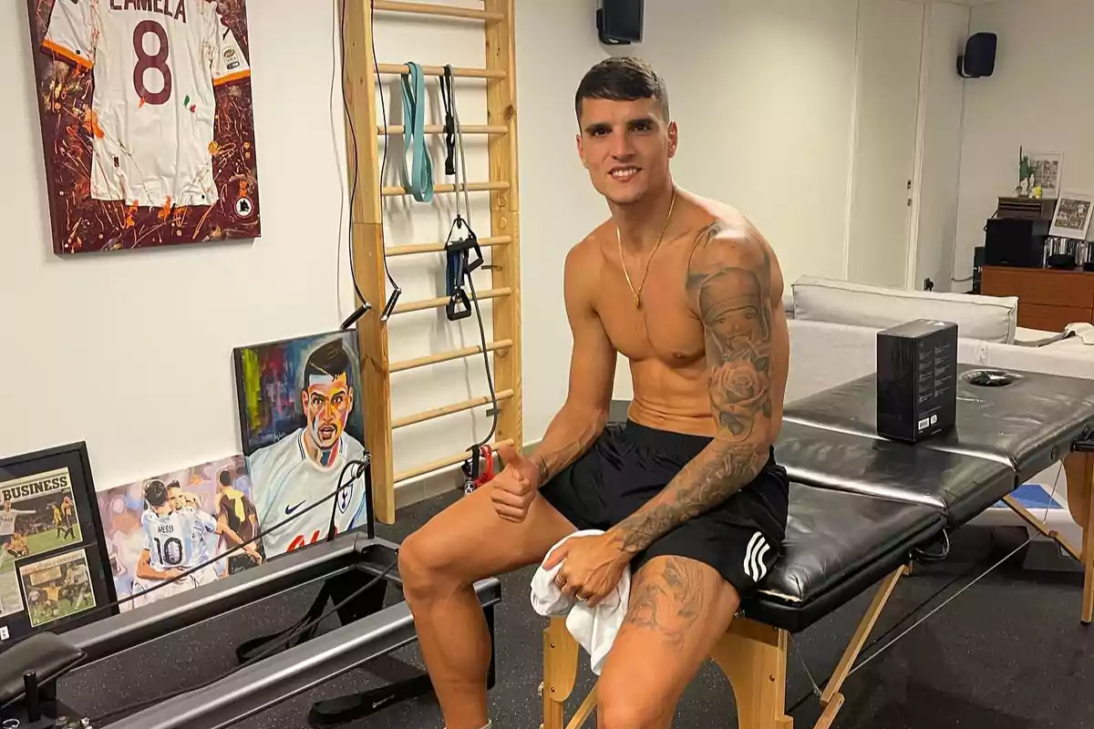 Hombre joven sin camiseta sentado en una camilla de fisioterapia mostrando el pulgar arriba en una sala decorada con cuadros, una camiseta enmarcada y equipo de ejercicio