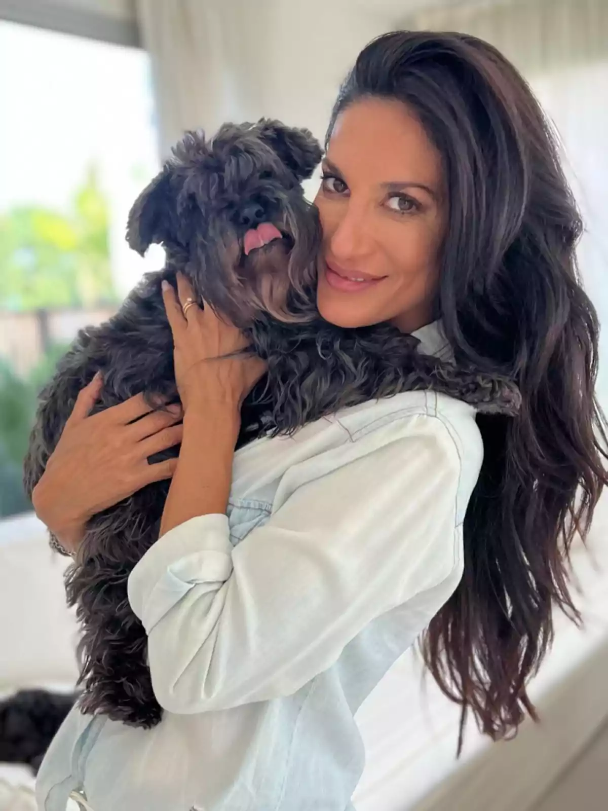 Mujer de cabello largo y oscuro sonríe mientras abraza a un perro pequeño de pelaje rizado y oscuro en una habitación iluminada Mujer de cabello largo y oscuro sonríe mientras abraza a un perro pequeño de pelaje rizado y oscuro en una habitación iluminada