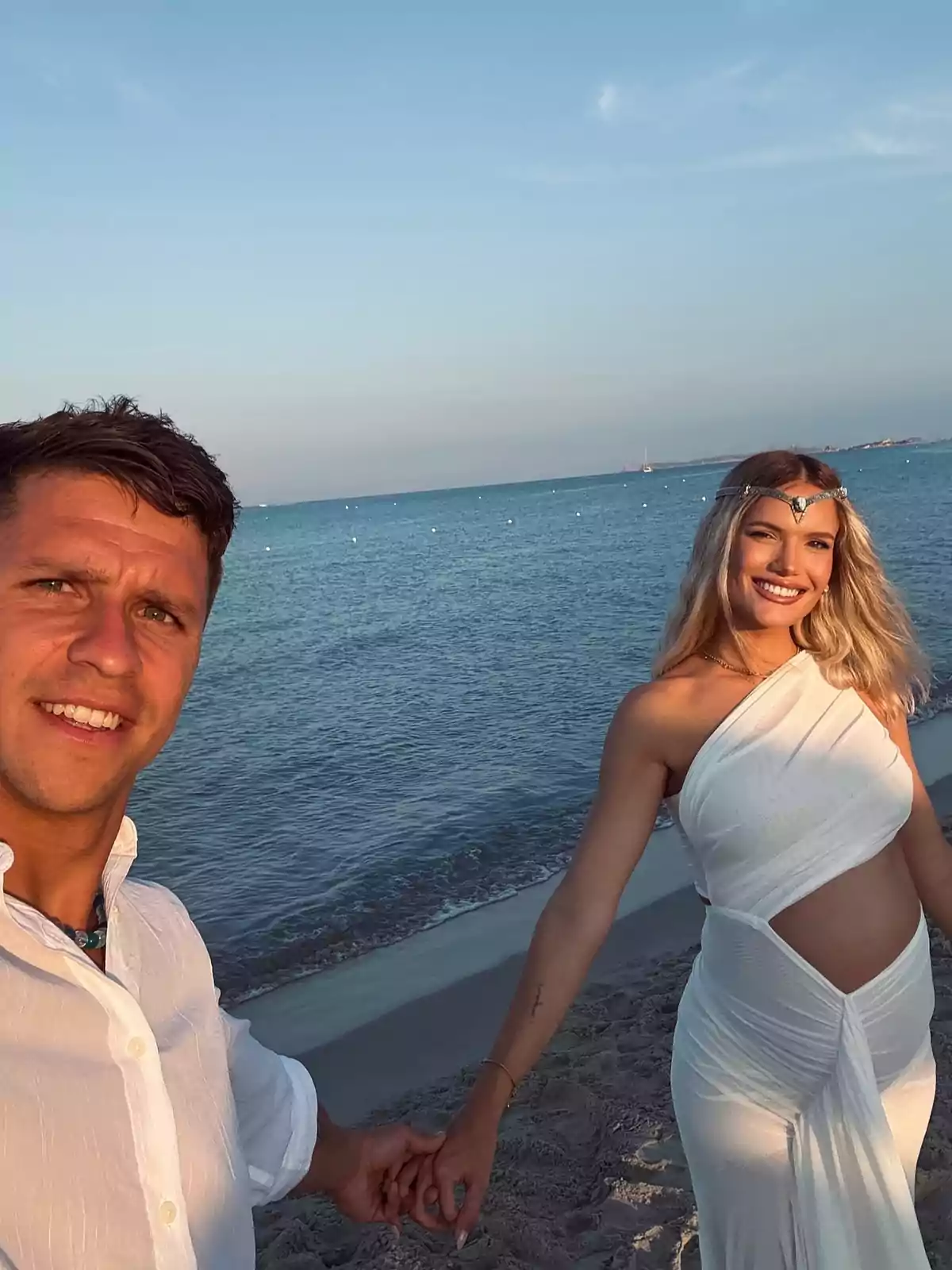 Smiling couple taking a selfie on the beach at sunset, both dressed in white and holding hands, with the sea in the background Smiling couple taking a selfie on the beach at sunset, both dressed in white and holding hands, with the sea in the background