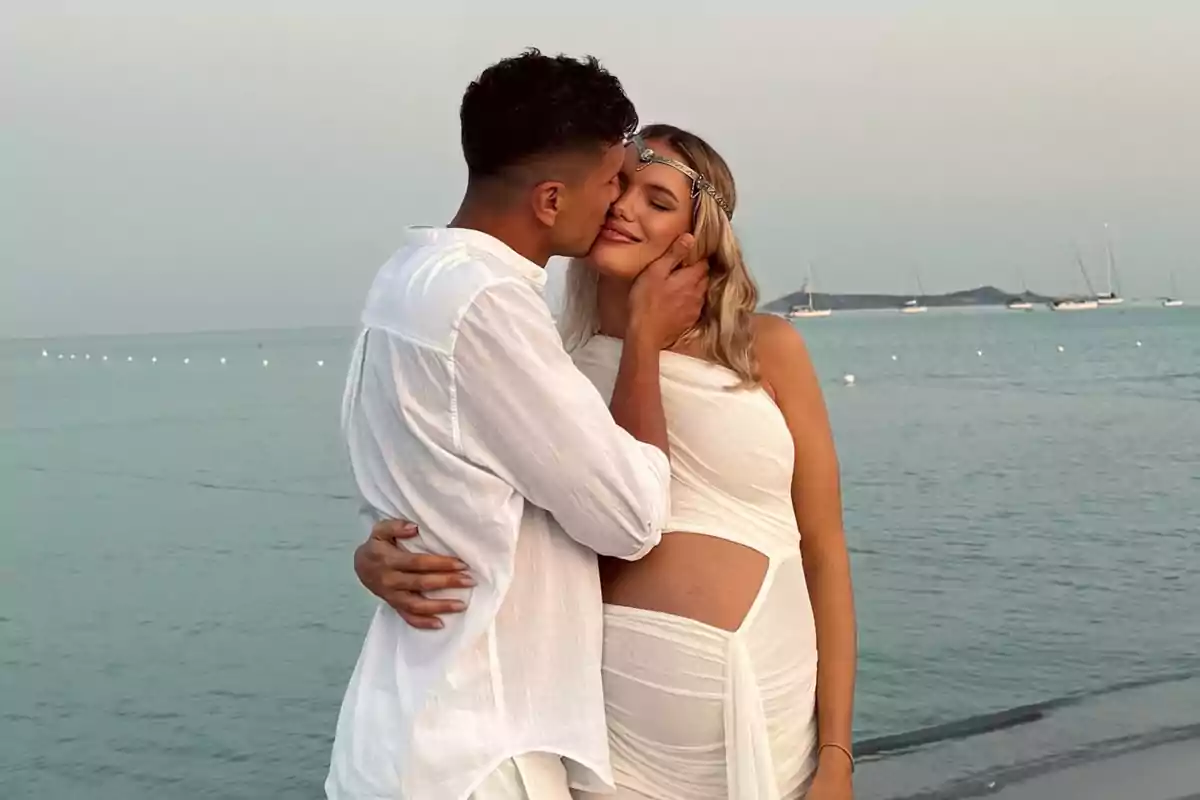 Couple hugging on the beach at sunset, both dressed in white, the pregnant woman smiles while the man kisses her on the cheek and boats can be seen in the sea in the background.