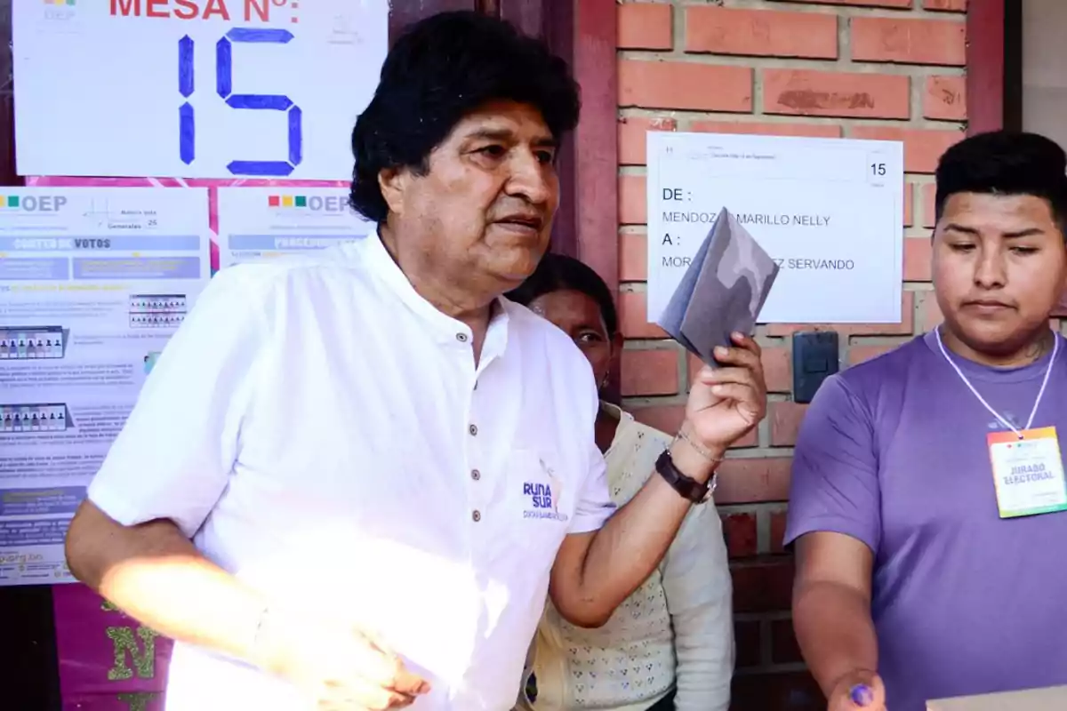 Hombre de cabello oscuro con camisa blanca sostiene un documento mientras se encuentra junto a un joven con credencial en un recinto de votación con carteles y un letrero de mesa número 15 al fondo Hombre de cabello oscuro con camisa blanca sostiene un documento mientras se encuentra junto a un joven con credencial en un recinto de votación con carteles y un letrero de mesa número 15 al fondo