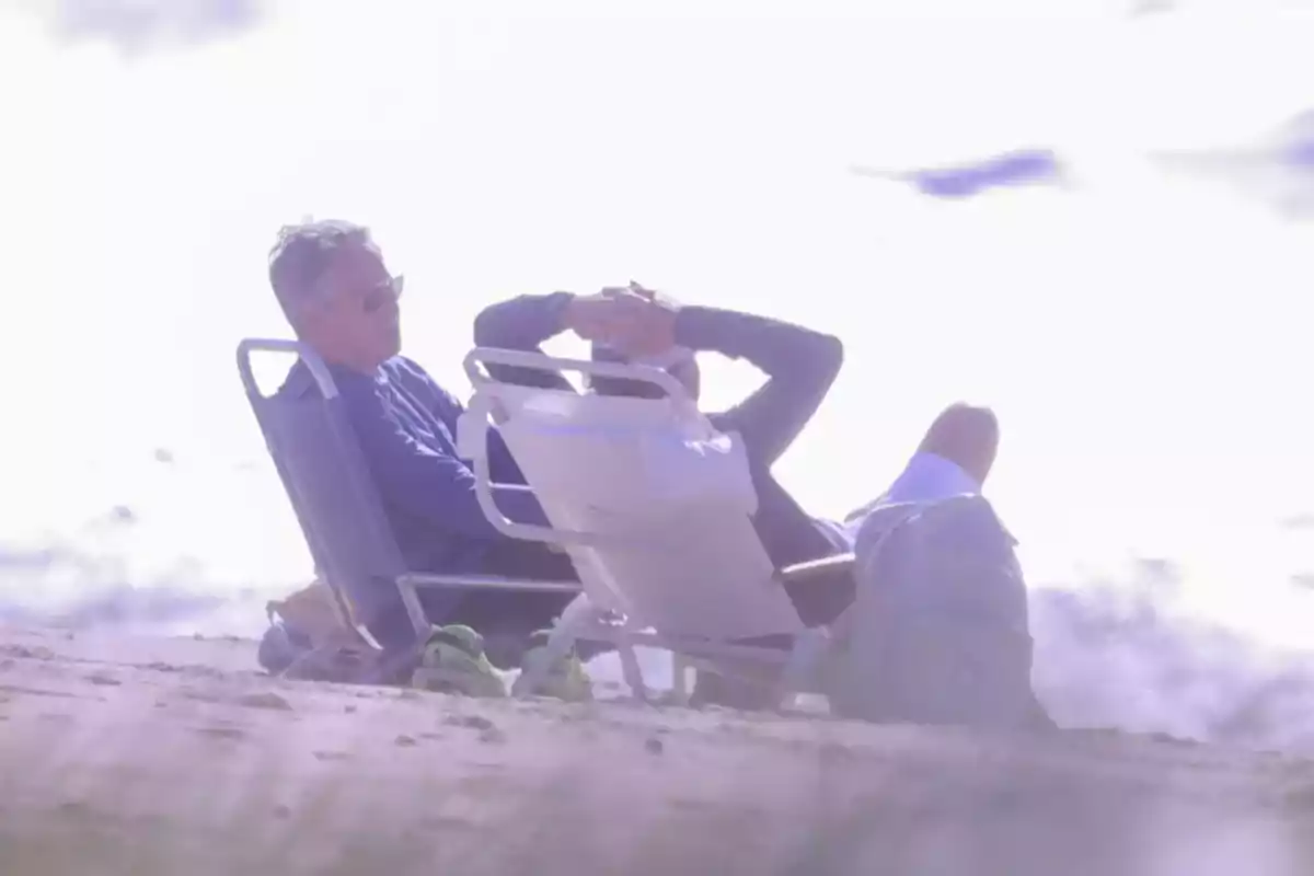 Hombre sentado en una silla de playa relajándose junto al mar con gafas de sol y ropa casual