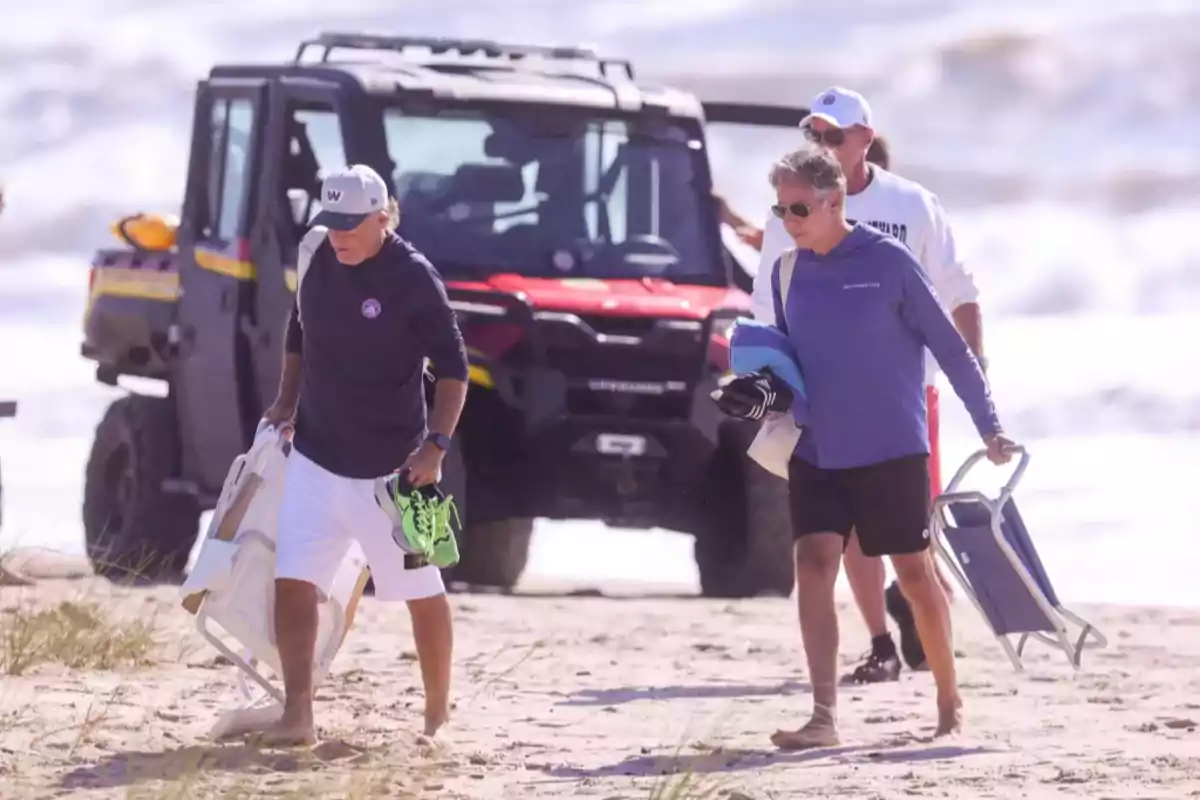 Tres hombres caminan descalzos por la playa llevando sillas plegables y objetos personales mientras un vehículo todoterreno está estacionado detrás de ellos Tres hombres caminan descalzos por la playa llevando sillas plegables y objetos personales mientras un vehículo todoterreno está estacionado detrás de ellos