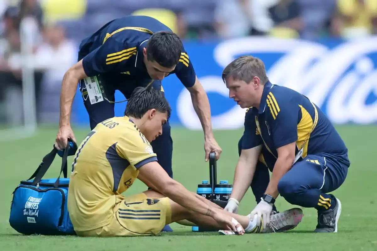A player sitting on the grass receives medical attention from two team members during a match. A player sitting on the grass receives medical attention from two team members during a match.