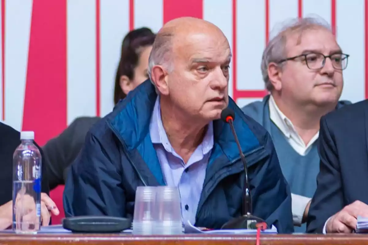 Bald man in a blue jacket speaking into a microphone at a table with other people.