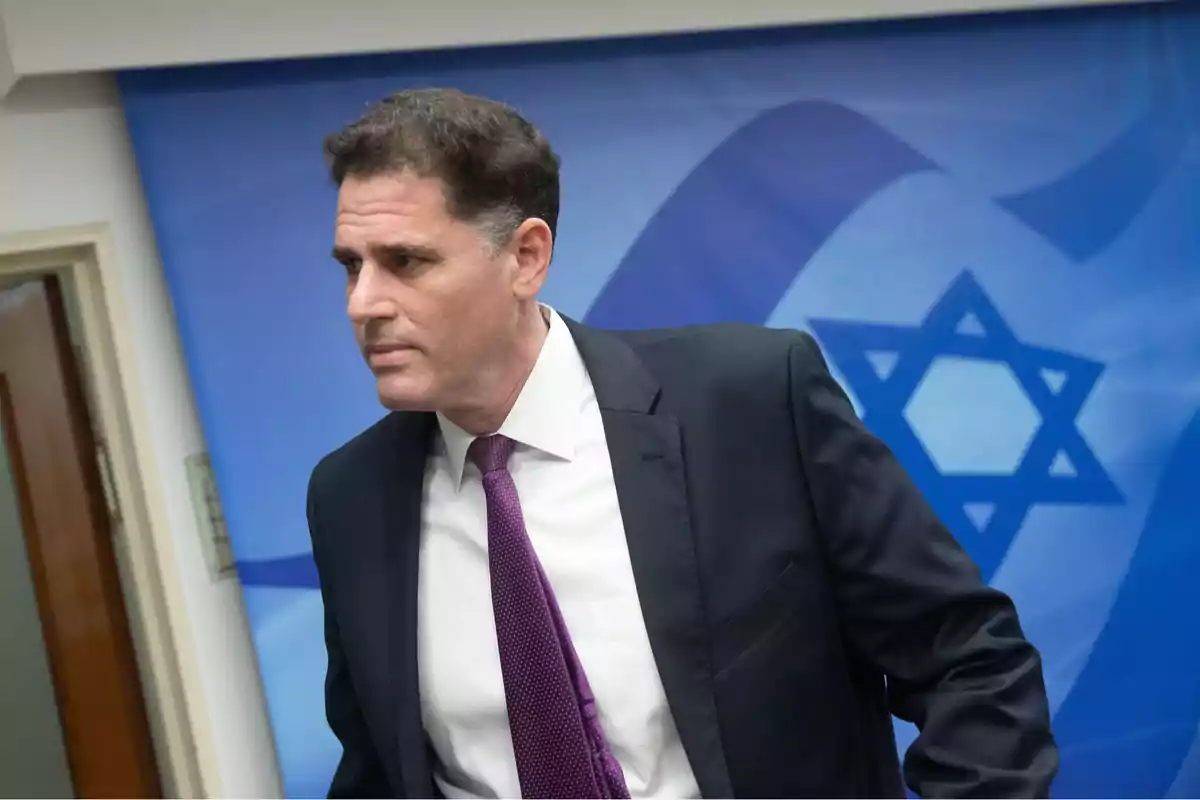 Man in a dark suit and purple tie in front of a blue flag with the Star of David