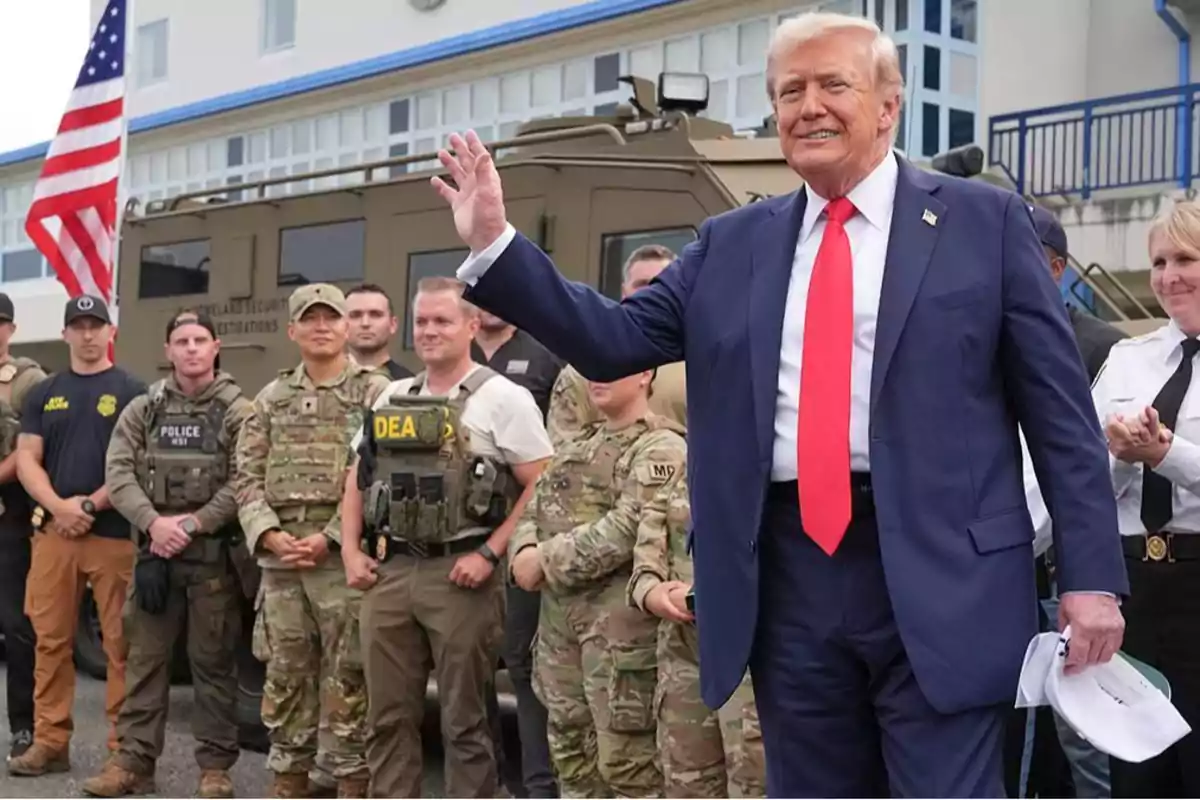 Hombre de traje azul y corbata roja saluda frente a un grupo de agentes y militares con uniforme, junto a un vehículo blindado y una bandera de Estados Unidos al fondo Hombre de traje azul y corbata roja saluda frente a un grupo de agentes y militares con uniforme, junto a un vehículo blindado y una bandera de Estados Unidos al fondo