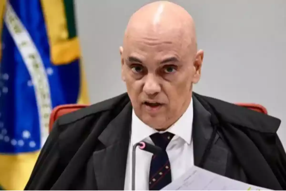 Bald man in a black robe and tie, sitting in front of a microphone with a Brazilian flag in the background Bald man in a black robe and tie, sitting in front of a microphone with a Brazilian flag in the background