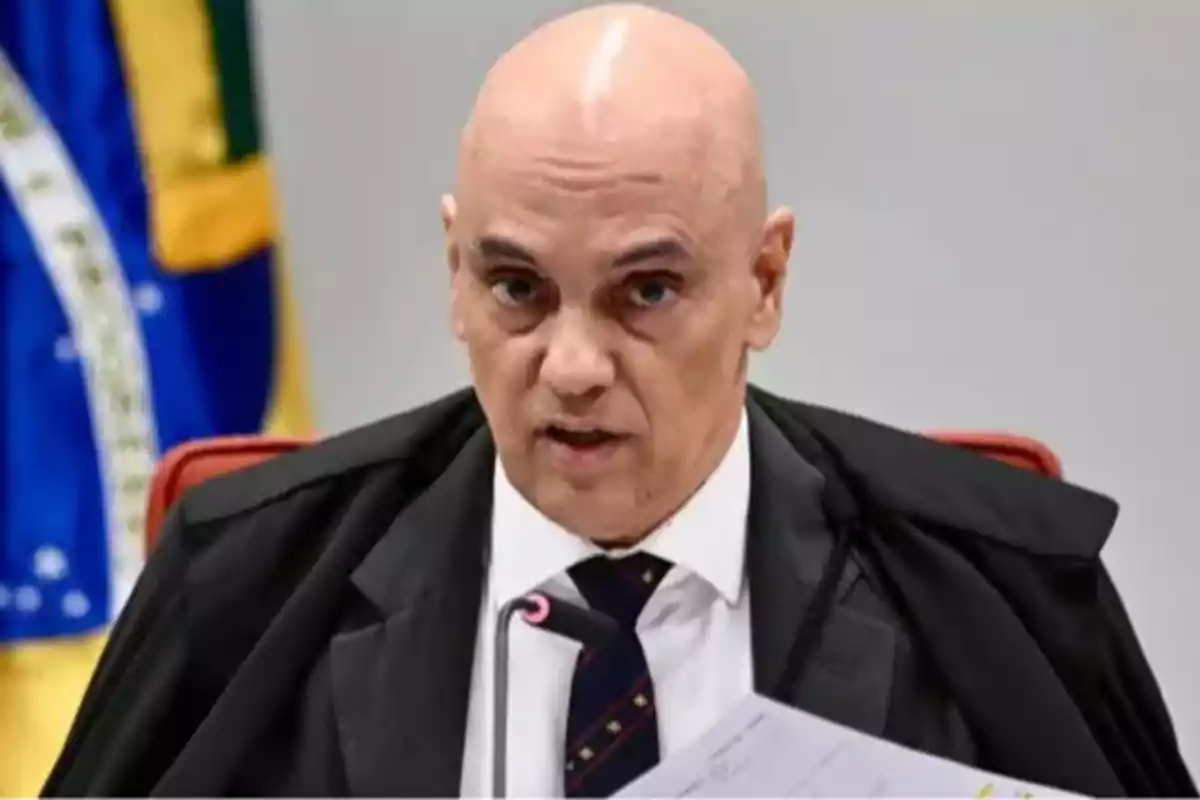 Bald man in a black robe and tie speaking in front of a microphone with a Brazilian flag in the background Bald man in a black robe and tie speaking in front of a microphone with a Brazilian flag in the background