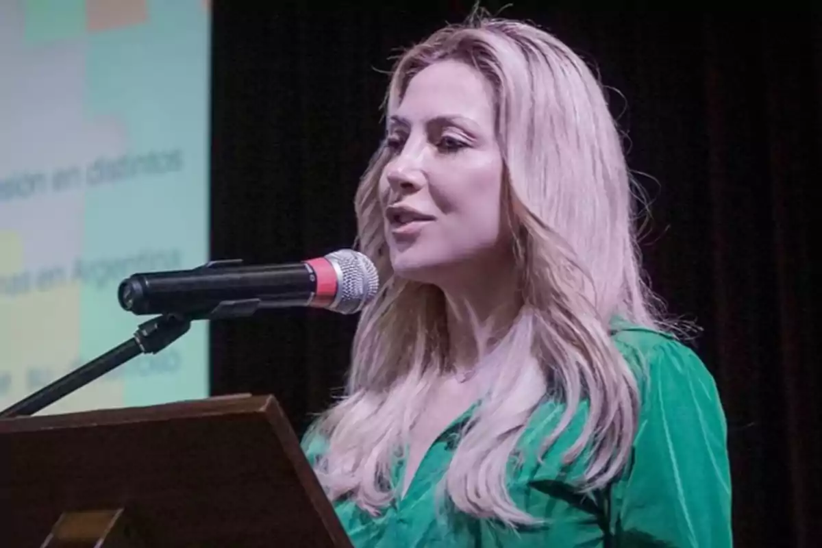 Blonde woman in a green blouse speaking in front of a microphone on a stage