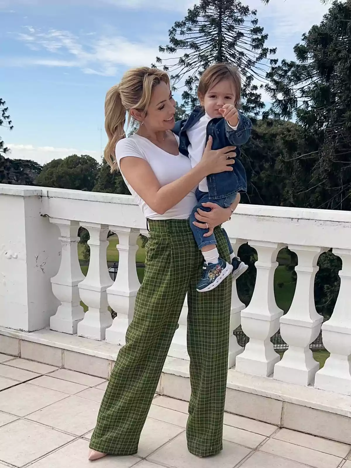 A smiling woman holds a small child in her arms while standing on a terrace with white railings and trees in the background. A smiling woman holds a small child in her arms while standing on a terrace with white railings and trees in the background.