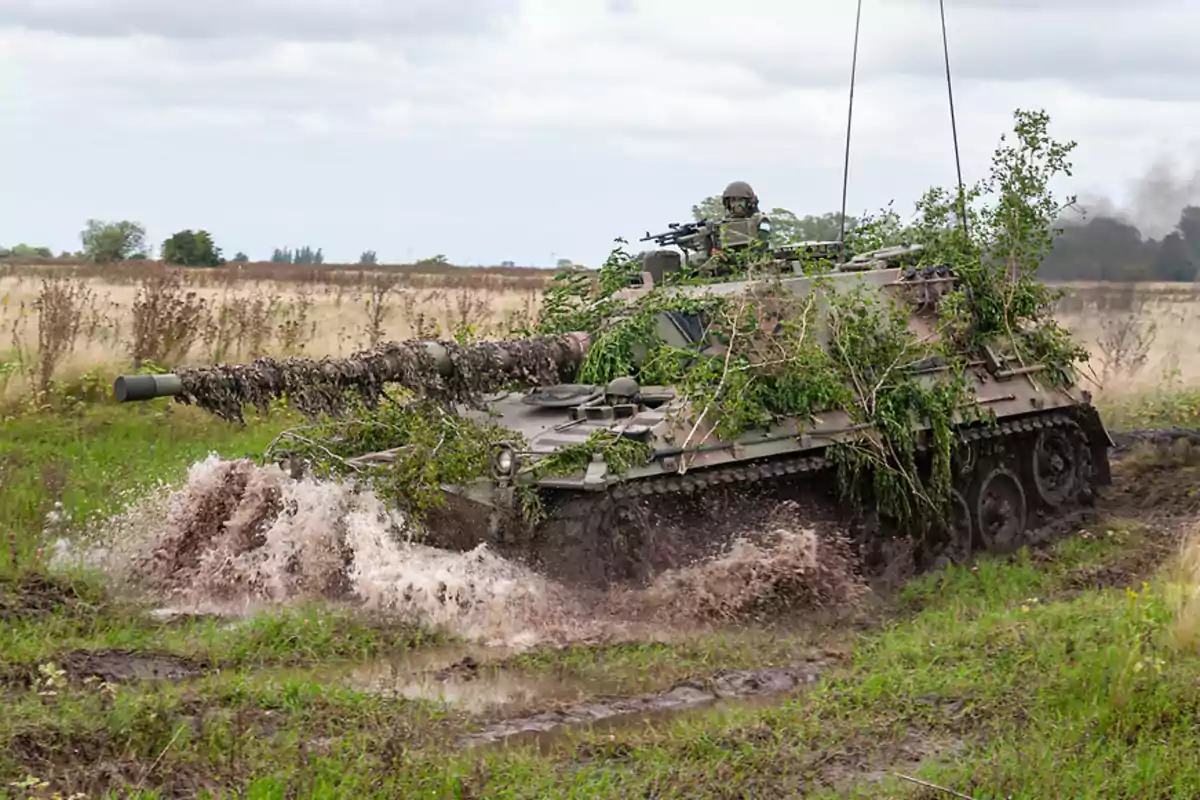 Camouflaged tank with branches advancing through muddy terrain and splashing water while a soldier peeks out from the hatch Camouflaged tank with branches advancing through muddy terrain and splashing water while a soldier peeks out from the hatch