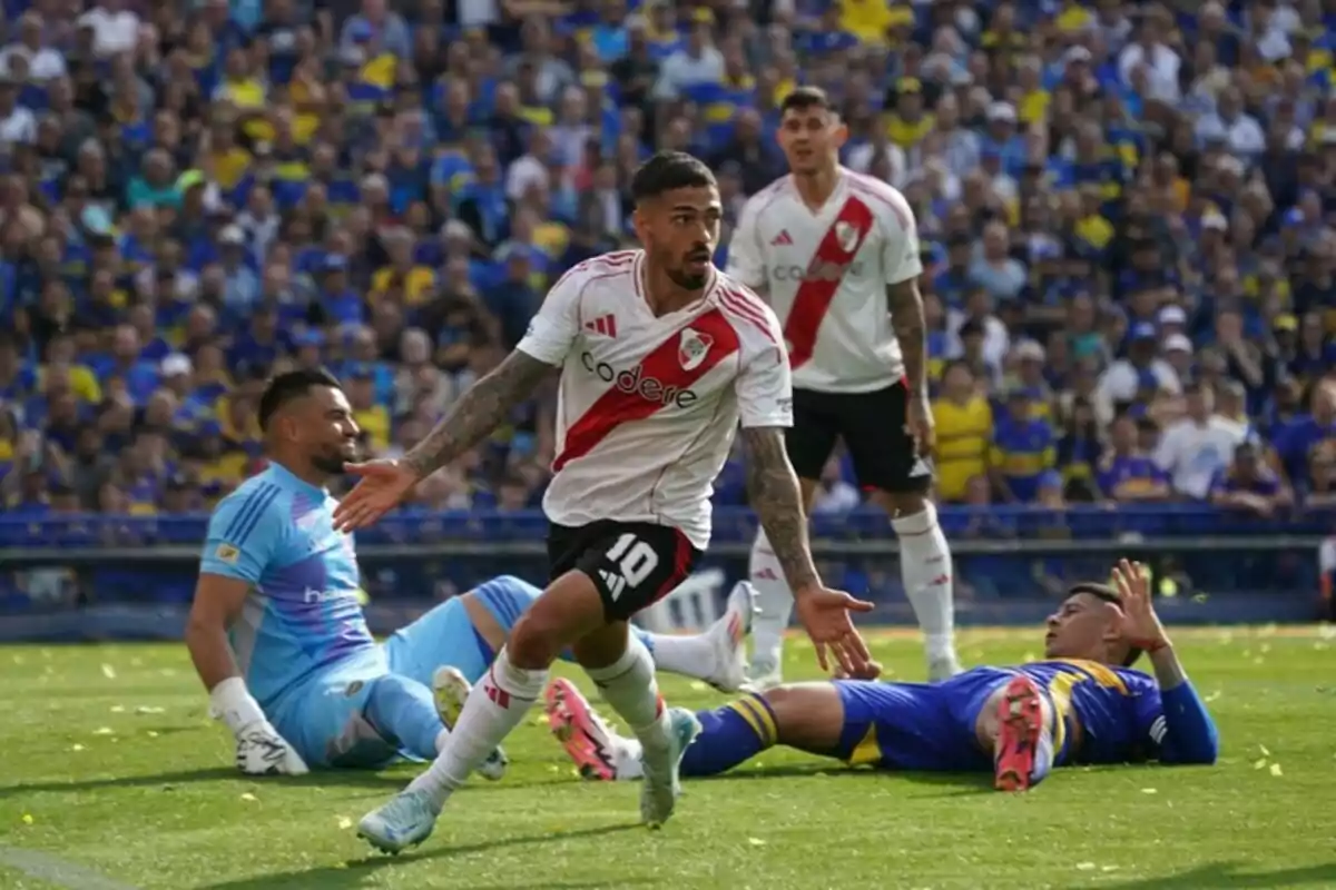 River Plate player celebrating a goal in front of Boca Juniors players during a soccer match with a stadium full of fans in the background River Plate player celebrating a goal in front of Boca Juniors players during a soccer match with a stadium full of fans in the background