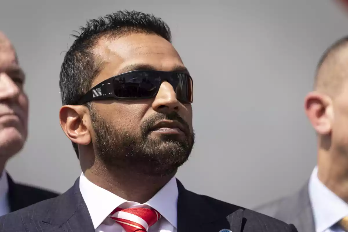 Man with a beard and dark sunglasses wearing a suit and red tie at an outdoor event