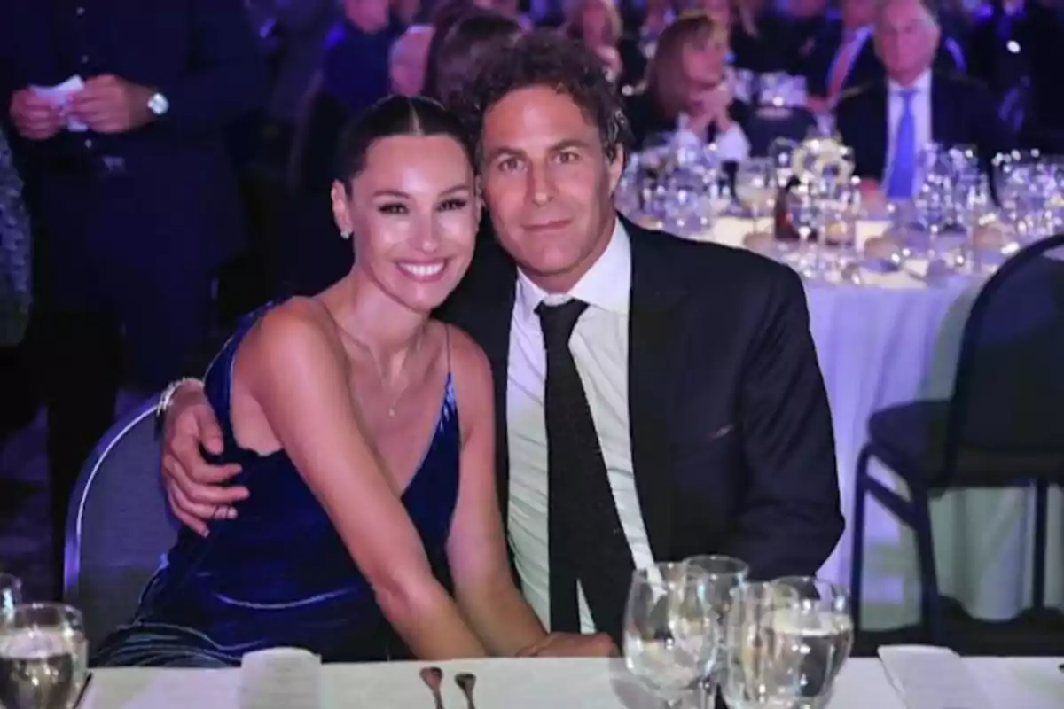 An elegantly dressed couple smiles while posing seated at a table during a formal event, surrounded by glasses and plates with other people in the background. An elegantly dressed couple smiles while posing seated at a table during a formal event, surrounded by glasses and plates with other people in the background.