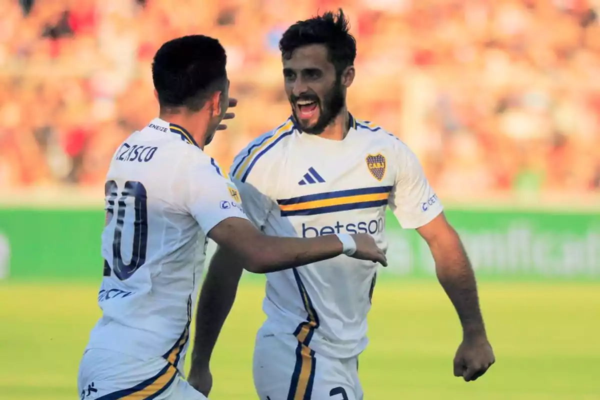 Two Boca Juniors players celebrate a goal in a soccer match, both wearing the team's white uniform and showing expressions of joy on the field. Two Boca Juniors players celebrate a goal in a soccer match, both wearing the team's white uniform and showing expressions of joy on the field.