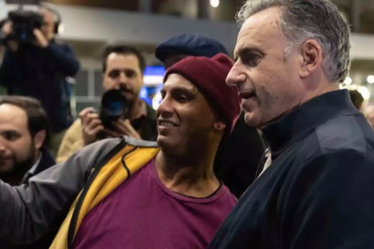 Two men pose for a selfie while several people with cameras surround them in a brightly lit indoor setting.