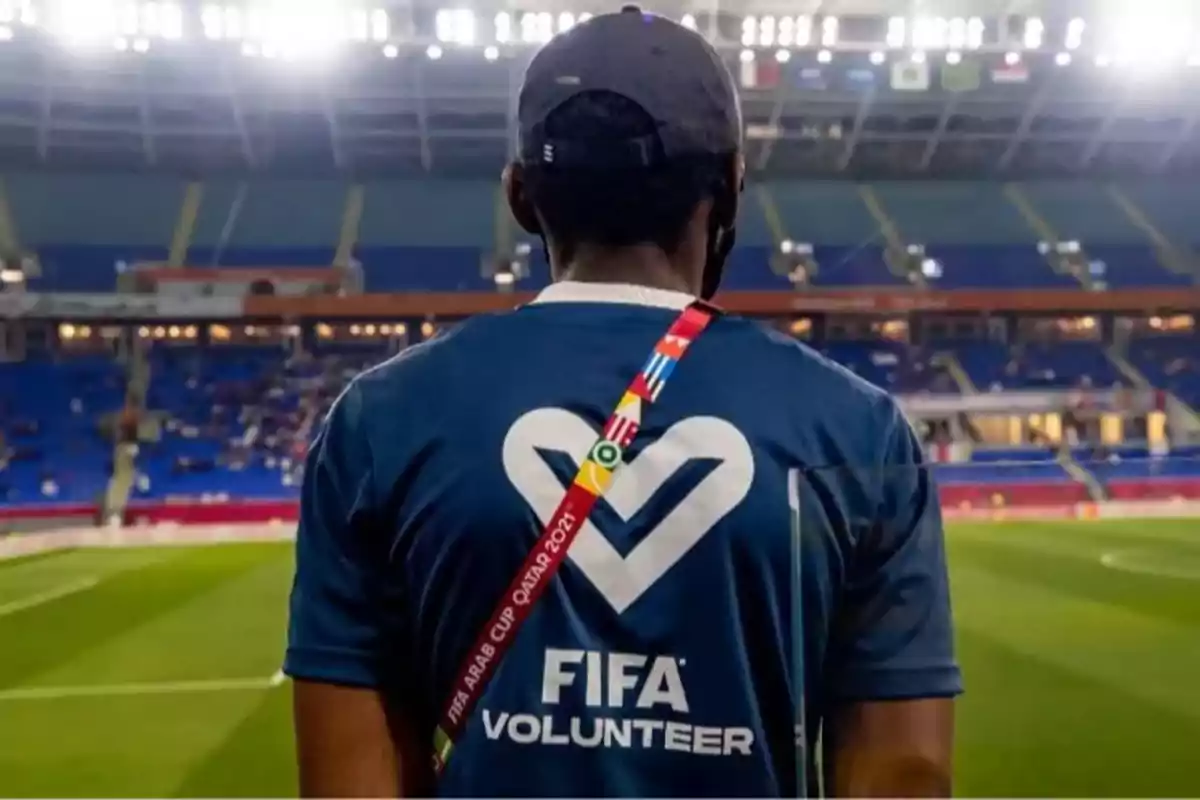 Persona de espaldas con camiseta de voluntario de la FIFA en un estadio de fútbol iluminado y con gradas parcialmente ocupadas