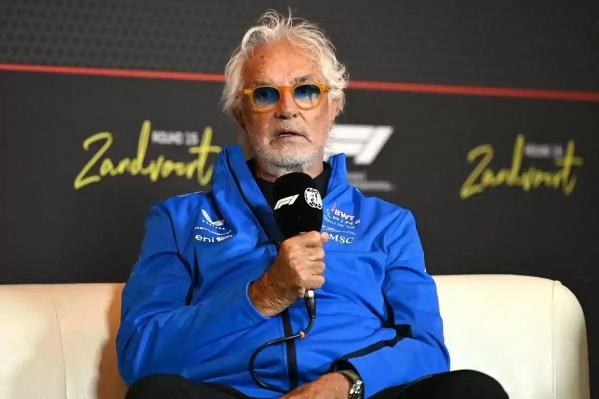 Gray-haired man with colorful glasses sitting on a white sofa wearing a blue jacket, holding a Formula 1 microphone during a press conference Gray-haired man with colorful glasses sitting on a white sofa wearing a blue jacket, holding a Formula 1 microphone during a press conference