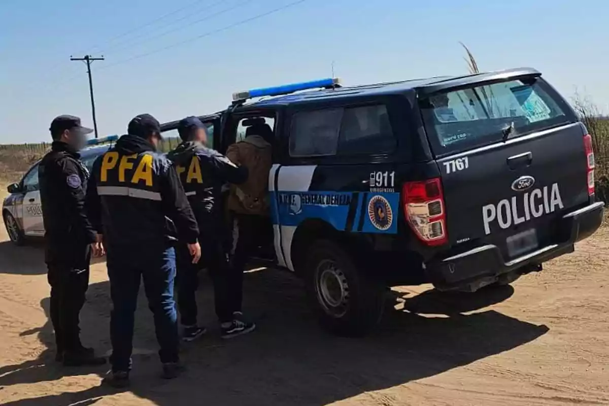 Several police officers detain a person and put them into a police truck on a dirt road during the day.