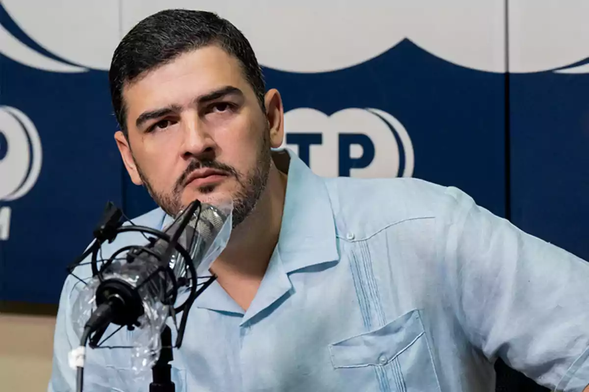 Hombre de cabello corto y barba usando una guayabera azul claro sentado frente a un micrófono en una cabina de radio