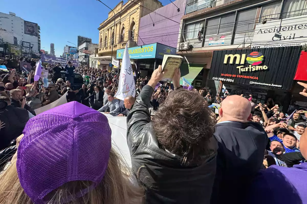 A crowd gathers on a shopping street during a demonstration while a person in the foreground holds up a book and others watch and take photos. A crowd gathers on a shopping street during a demonstration while a person in the foreground holds up a book and others watch and take photos.