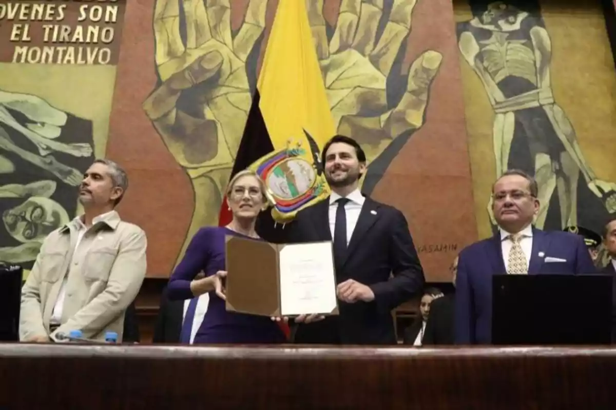 Four people pose at an official event in front of a mural and an Ecuadorian flag, one of them holding an open document.