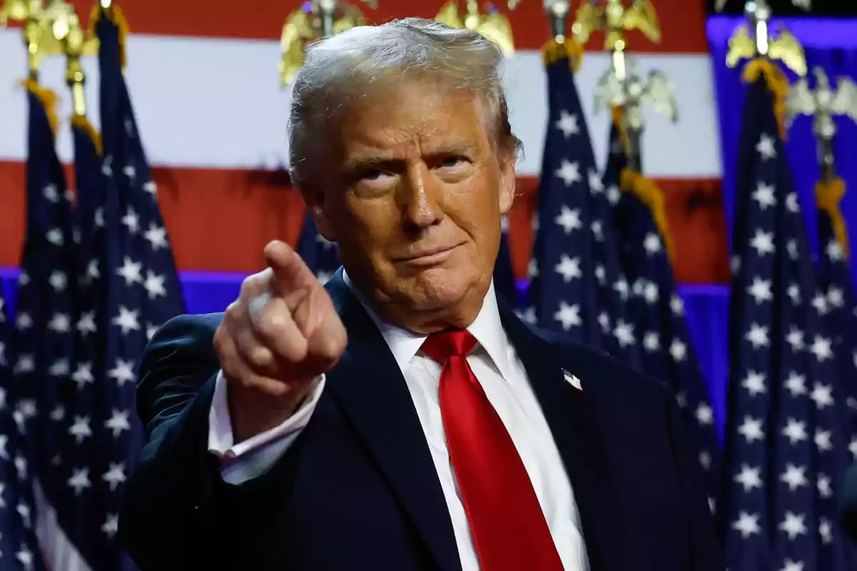 Blond-haired man in a dark suit and red tie pointing forward with several American flags in the background