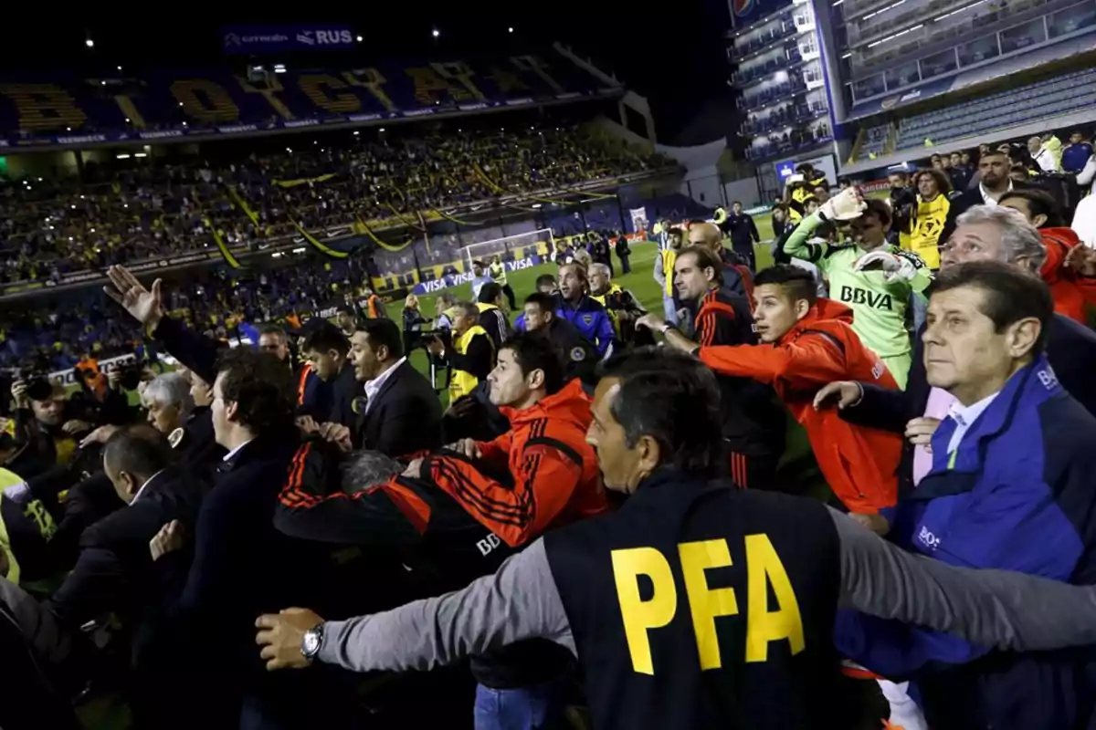 A group of people, including players, security personnel, and officials, are on the field of a soccer stadium packed with fans, in a tense and chaotic atmosphere. A group of people, including players, security personnel, and officials, are on the field of a soccer stadium packed with fans, in a tense and chaotic atmosphere.