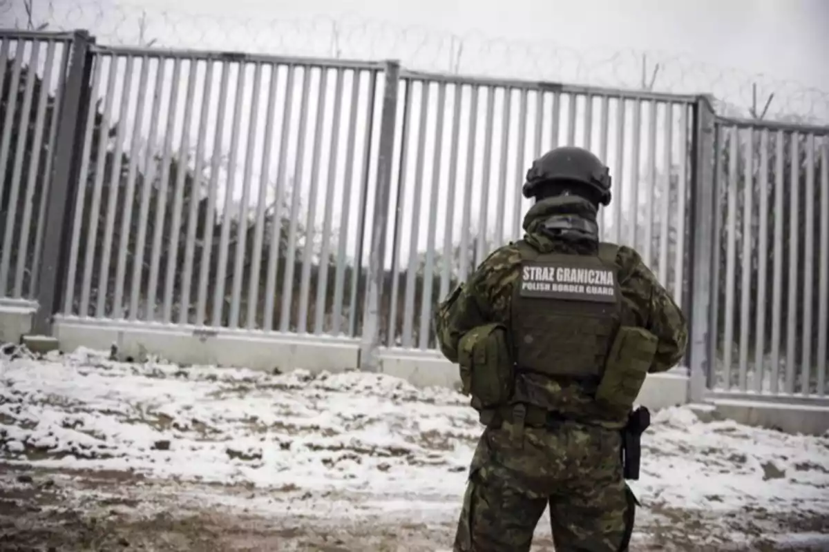 Un guardia fronterizo polaco con uniforme militar y casco observa una valla alta de metal en una zona nevada.