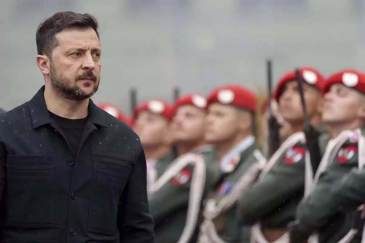 Un hombre de barba y chaqueta oscura está de pie al frente mientras un grupo de soldados con boinas rojas y uniformes verdes permanece alineado al fondo
