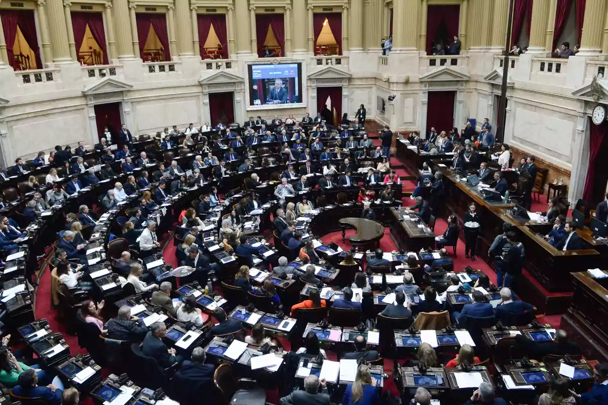 Panoramic view of a parliamentary session with numerous legislators seated at their desks inside a chamber with classical architecture and red curtains