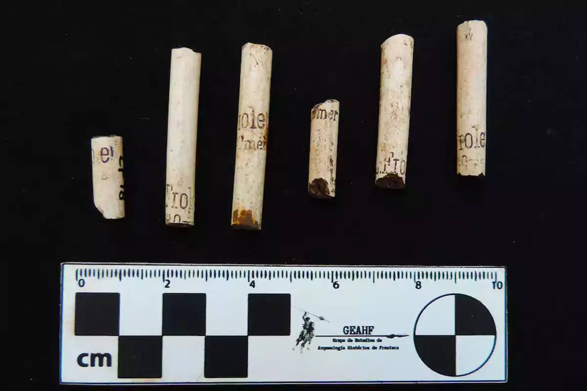 Siete fragmentos de pipas de cerámica alineados sobre una superficie negra junto a una regla métrica para escala Siete fragmentos de pipas de cerámica alineados sobre una superficie negra junto a una regla métrica para escala