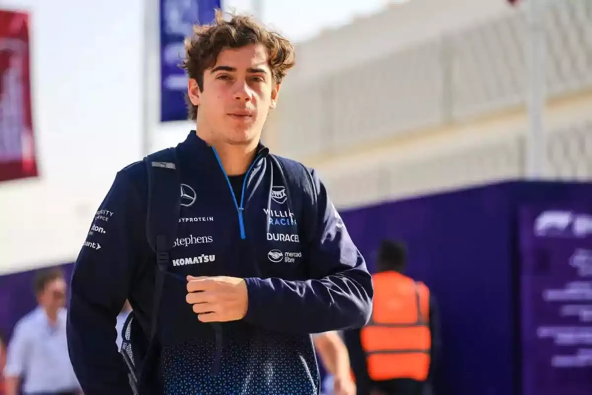 Young man wearing a Williams Racing blue jacket and a backpack walking in an outdoor sports environment Young man wearing a Williams Racing blue jacket and a backpack walking in an outdoor sports environment