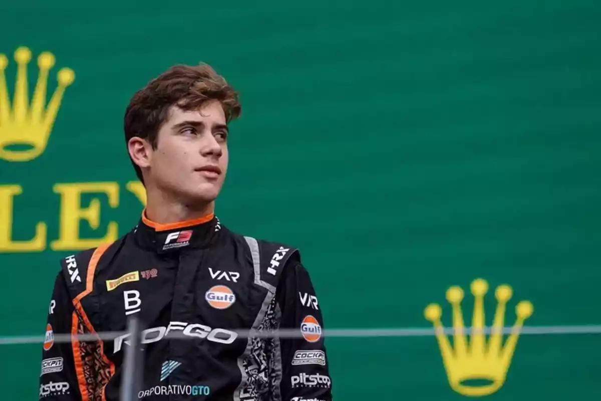 Young motorsport driver in a black and orange racing suit posing in front of a green background with yellow logos Young motorsport driver in a black and orange racing suit posing in front of a green background with yellow logos
