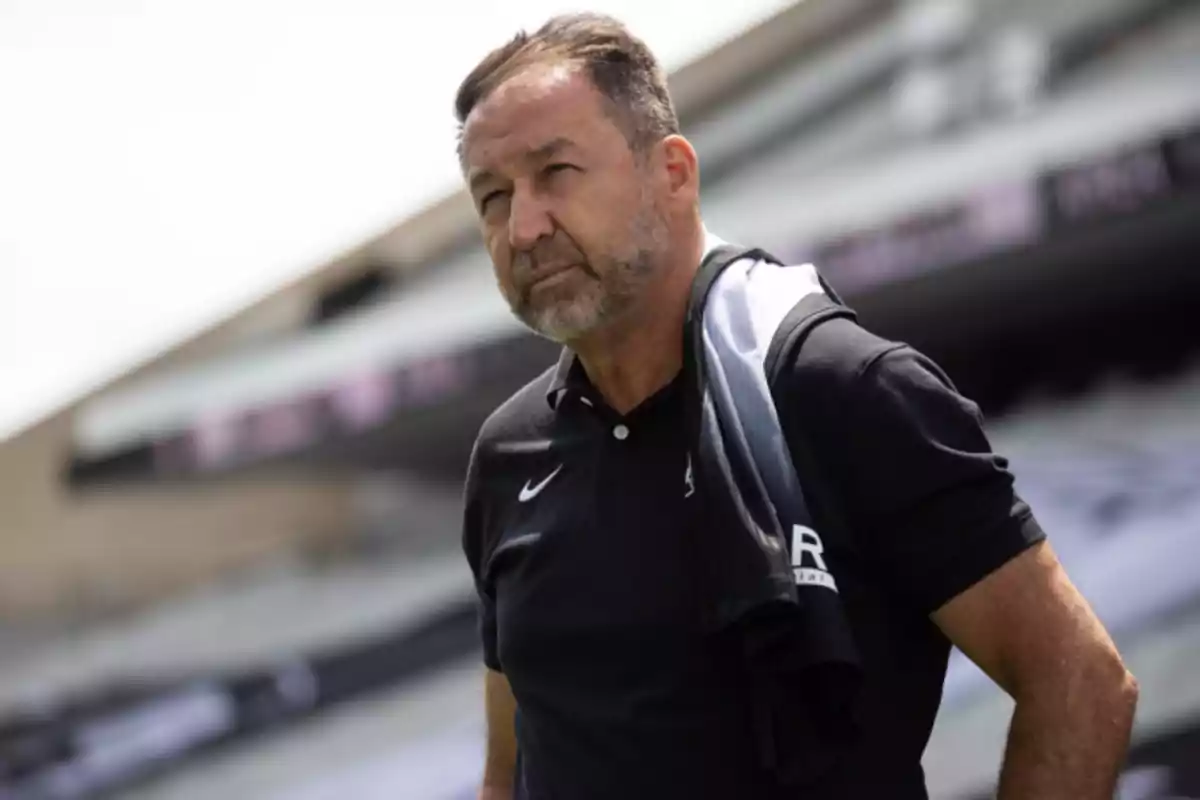 Middle-aged man with a beard and short hair wears a black polo and has a garment over his shoulder in a sports stadium.