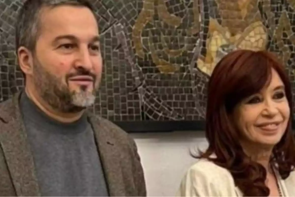 Dos personas posan sonrientes frente a un mural de mosaicos en tonos oscuros y dorados