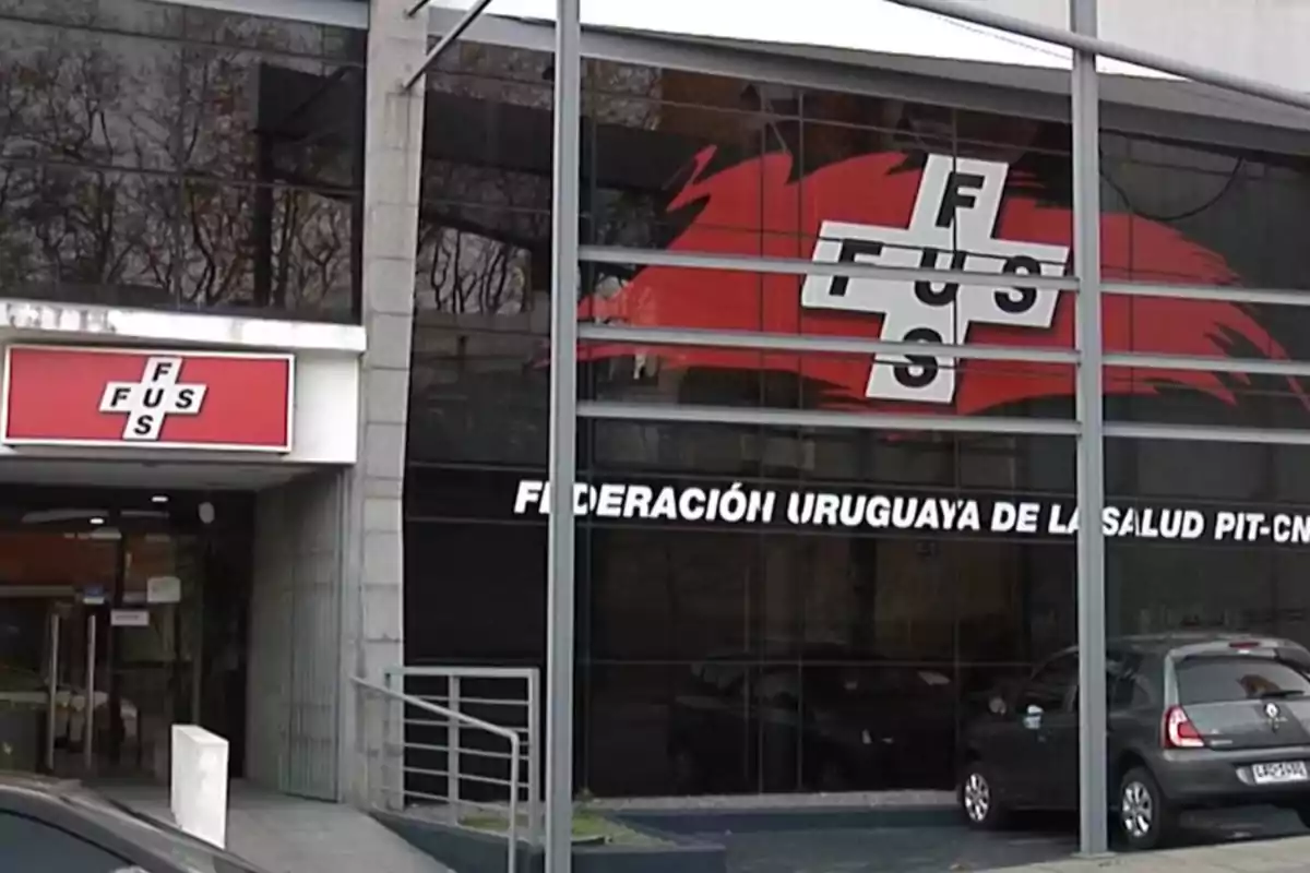 Fachada de un edificio con el logo de la Federación Uruguaya de la Salud y autos estacionados frente a la entrada Fachada de un edificio con el logo de la Federación Uruguaya de la Salud y autos estacionados frente a la entrada